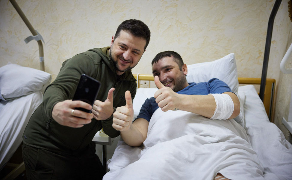 Kuvassa Ukrainan presidentti Volodymir Zelenskyi (vasemmalla) vierailee sairaalassa. Hän ottaa selfien yhdessä haavoittuneen sotilaan kanssa.