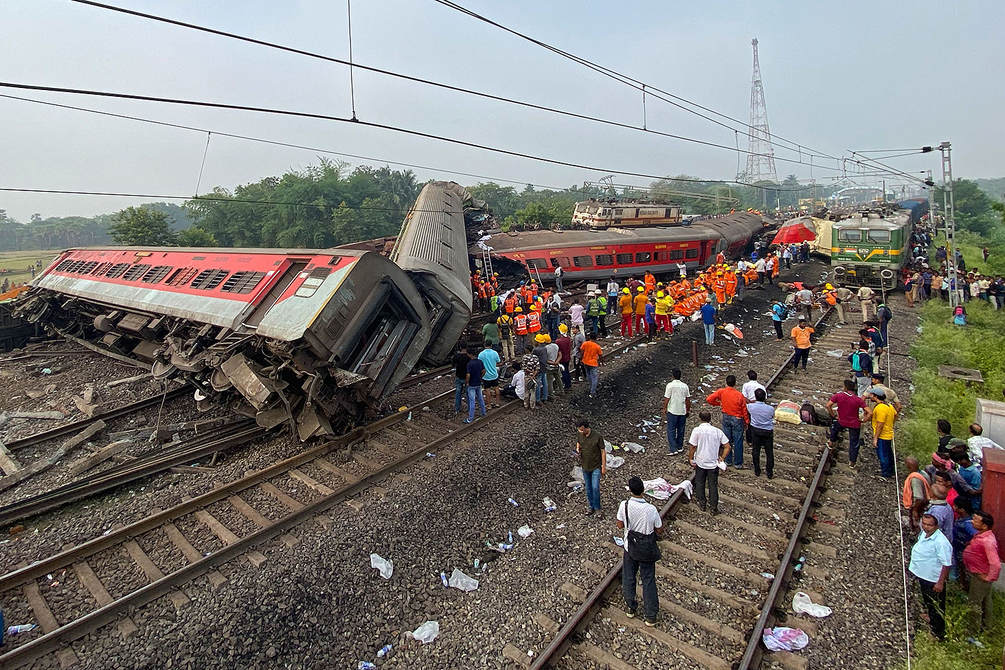 Ihmiset ovat kokoontuneet kaatuneiden junavaunujen ympärille. Kuvassa näkyy myös paljon pelastustyöntekijiöitä, joilla on kirkkaan oranssit vaatteet ja kypärät. Osa junavaunuista on poikittain junaradalla kaatuneena. Kuva on otettu lauantaina 3. kesäkuuta Balasoren kaupungissa Itä-Intiassa.