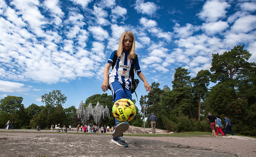 Lapsi nostaa jalallaan jalkapalloa. Hänellä on päällä pelivaatteet ja selässä reppu. Taustalla kaukana Sibelius-monumentti ja ihmisiä katsomassa monumenttia eli veistosta.