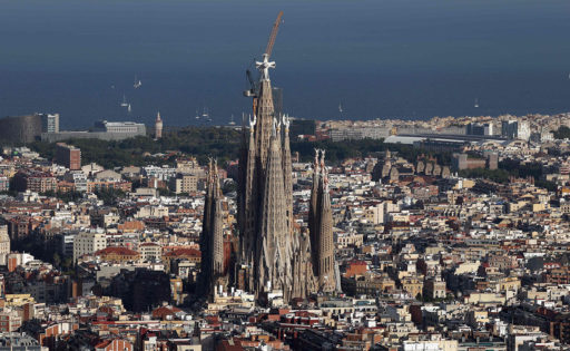 Sagrada familia -kirkko Barcelonassa. Kirkko on kuvattu ilmasta ja kuvassa näkyy kaupungin rakennuksia ja merta.