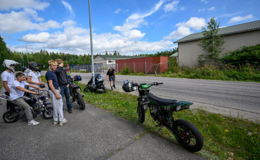 Nuoria mopoilijoita ja mopoja tien vieressä jalkakäytävällä. Autotien ja jalkakäytävän välissä on nurmikkoa.